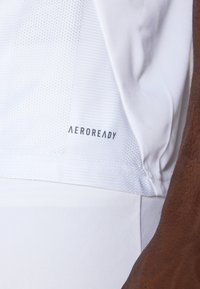 Hvid atletisk skjorte med åndbart mesh-textur, prydet med "AEROREADY" branding på siden, glat stof og en tætsiddende design.
