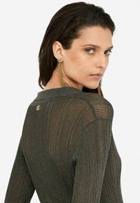 Grüne gerippte Langarmstrickjacke mit metallischen Fäden, rundem Halsausschnitt und kleinem Logo-Detail auf dem Rücken. Das Model trägt Quastenohrringe.