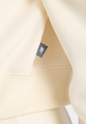 Sweatshirt de couleur crème avec une texture douce et côtelée. Présente une petite étiquette grise avec le logo sur la couture, mettant en avant un design simple et une coupe décontractée.