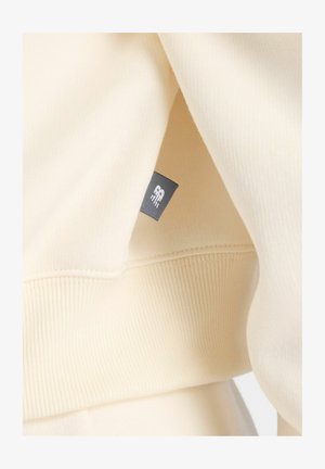 Sweatshirt de couleur crème avec une texture douce et côtelée. Présente une petite étiquette grise avec le logo sur la couture, mettant en avant un design simple et une coupe décontractée.