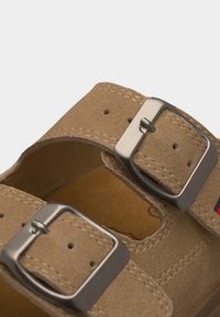 Κοντινό πλάνο σε λουριά δερμάτινου σανδαλιού ταμπά με ασημένιες μεταλλικές αγκράφες και λεπτομέρειες ραφής σε ανοιχτό φόντο.