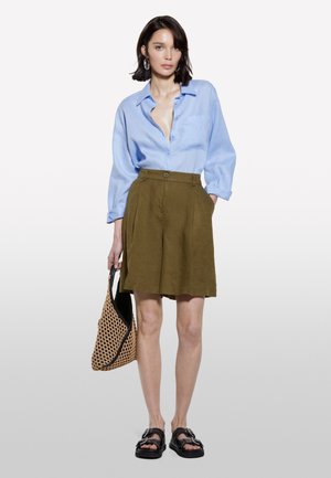 Chemise à manches longues bleu clair, short plissé vert olive, sandales noires, portant un sac en toile tressée. Tissu doux ; coupe décontractée.