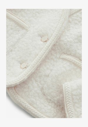 Vitt ylleblandat överkast med texturerad, mjuk yta. Har flätad kant och runda metallknappar. Närbild visar sömmönster.