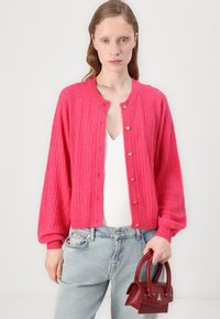 Hett rosa stickad cardigan med rund halsringning, ribbat mönster och guldhjärtknappar. Matcher med ljusblå denim och en liten bordeauxhandväska.