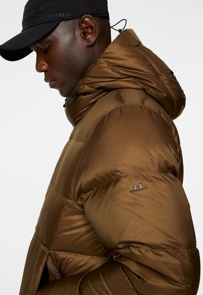 Brun puffjacka med avtagbar huva, som har ett vadderat mönster och en broderad logotyp på ärmen. Svart keps kompletterar outfiten.