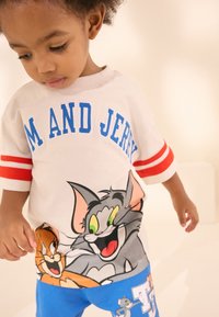 Vit T-shirt med rödrandiga ärmar, med en grafisk bild av tecknade figurerna Tom och Jerry, kombinerat med blå shorts med figurgrafik.
