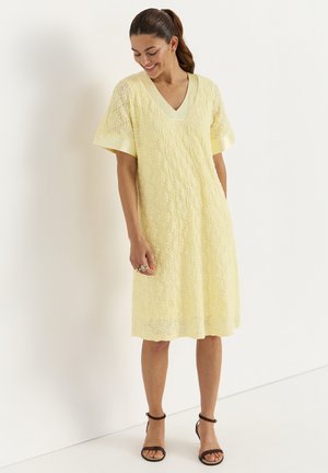 Vrouw glimlachend en omlaag kijkend, gekleed in een knie-lange gele kanten jurk met V-hals en korte mouwen, gecombineerd met zwarte enkellaarsjes met enkelbandje.