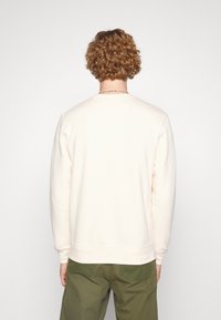 Světle růžová mikina z měkkého materiálu, s kulatým výstřihem, žebrovanými manžetami a volným střihem. Nošena s olivově zelenými kalhotami.