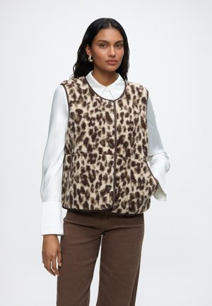 Femme portant une veste sans manches en polaire imprimé léopard marron et crème sur une chemise blanche colletée avec un pantalon marron, debout devant un fond uni.