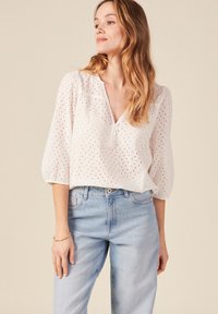Weiße Baumwollbluse mit Lochmuster, V-Ausschnitt und 3/4 Puffärmeln. Kombiniert mit hellblauen Jeans. Einfaches und atmungsaktives Design.