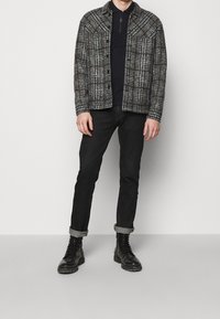 Tkaný kostkovaný kabát v šedé, černé a hnědé barvě; zkombinovaný s černým polo tričkem. Úzké černé džínsy a robustní černé šněrovací boty dotvářejí vzhled.