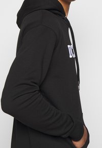 Sudadera negra de tela suave, con un logotipo bordado en blanco en el pecho, con cordones y detalle de bolsillo frontal.