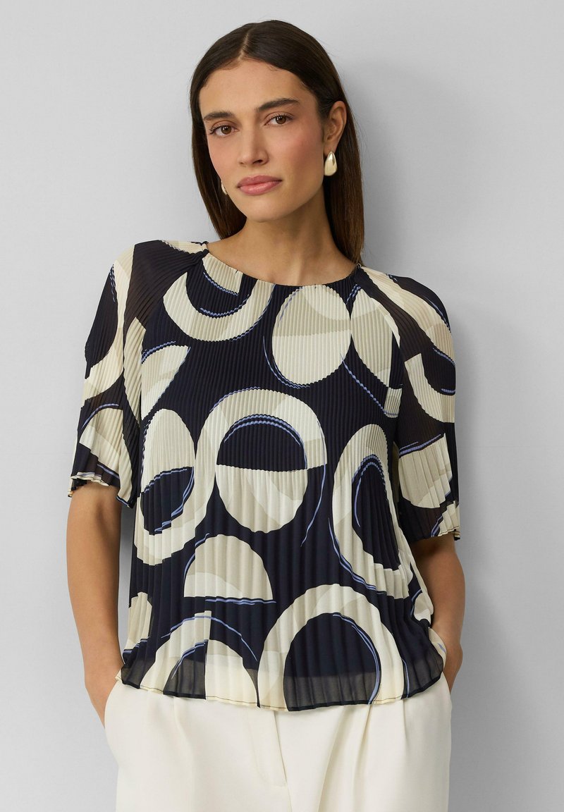Blouse plissée bleu marine avec des motifs circulaires beiges et blancs, dotée de manches courtes et d'un col rond. Texture lisse, coupe décontractée.