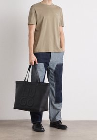 Mand iført patchwork denim bukser, en beige T-shirt, sorte sko og holder en stor sort tote bag med "HUGO" præget.