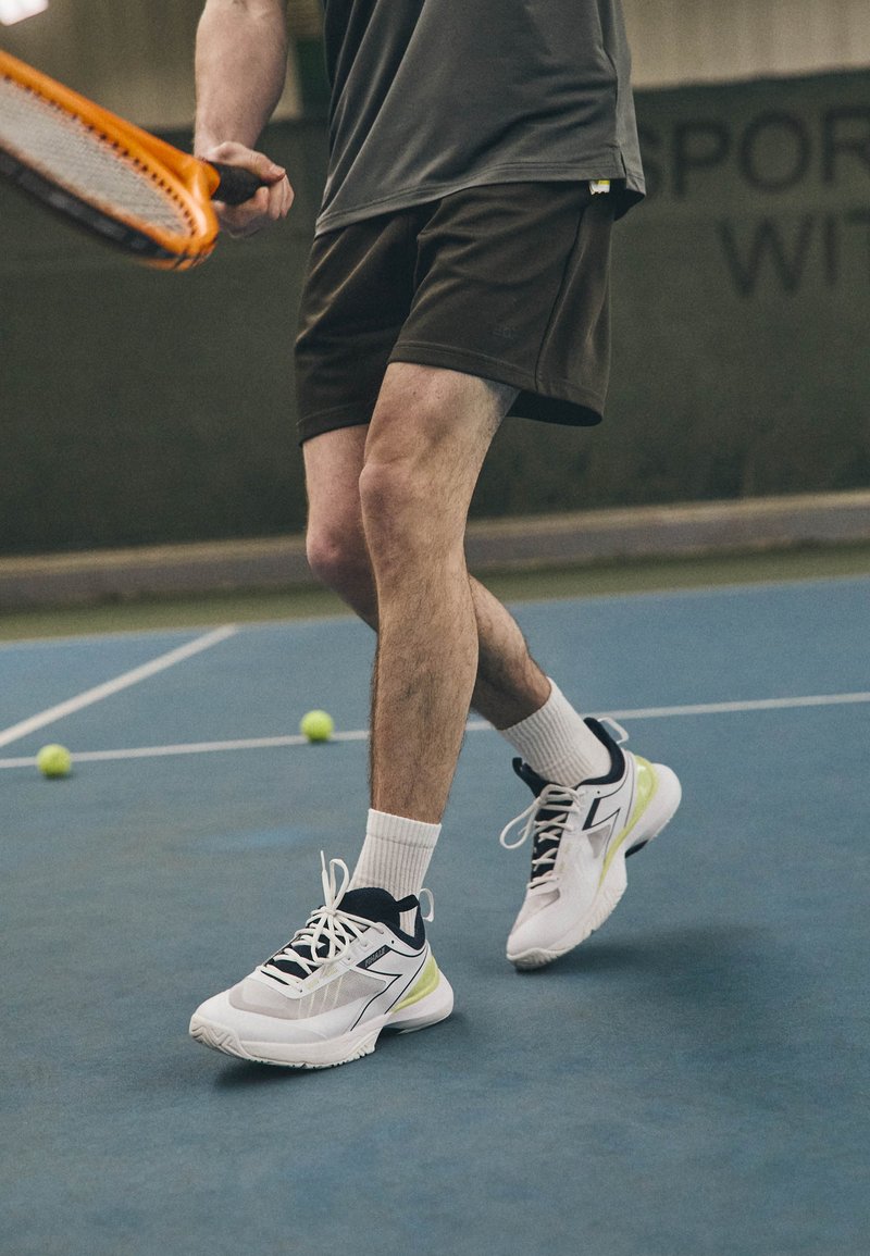 Vita atletiska skor med svarta och gula detaljer, bärs med vita strumpor, ihopparade med mörka shorts, på en blå tenniscourt med gröna bollar.