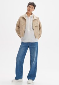 Beige Bomberjacke mit hohem Kragen und zwei Fronttaschen, kombiniert mit einem weißen Hemd und weit geschnittenen blauen Jeans. Weiße Sneakers.