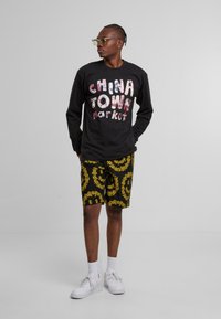 Svart langermet skjorte med "CHINA TOWN Market" i flerfarget tekst, kombinert med svarte shorts med et gult blomstermønster i spiral. Hvite joggesko.