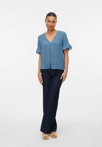 Blusa corta a maniche corte di colore azzurro chiaro con scollatura a V e dettagli con bottoni sul davanti, abbinata a jeans a gamba larga in denim scuro e tacchi aperti gialli.