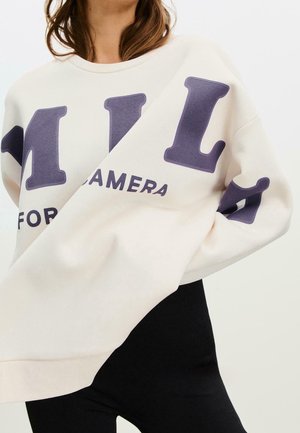 Femme portant un sweat-shirt blanc surdimensionné avec de grandes lettres violet foncé et des leggings noirs, fond blanc uni.