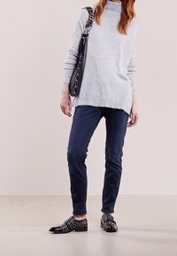 Grå polotröja, marinblå slim-fit byxor, svarta dekorerade skor och en svart nitaraxelväska. Släta texturer och minimalistisk design.