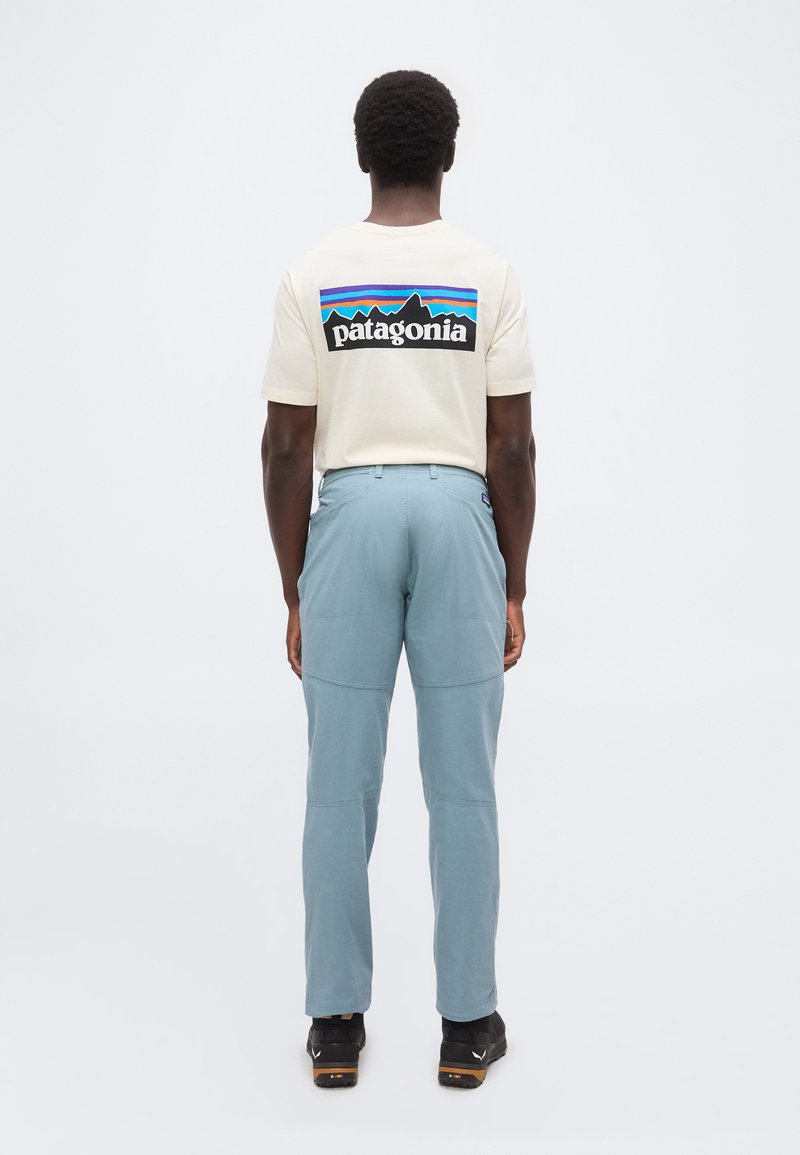 Ein Mann, der von hinten zu sehen ist, trägt ein cremefarbenes Patagonia T-Shirt mit Logodruck und hellblaue Hosen sowie schwarze Wanderschuhe.