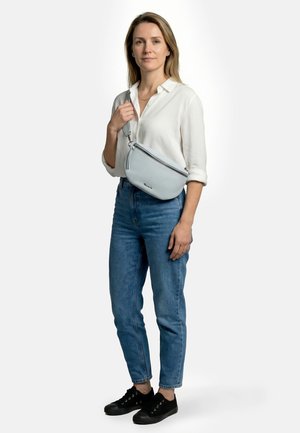 Mujer de pie mirando hacia adelante, vestida con blusa blanca, jeans azules, zapatillas negras y un bolso cruzado azul claro sobre el pecho.