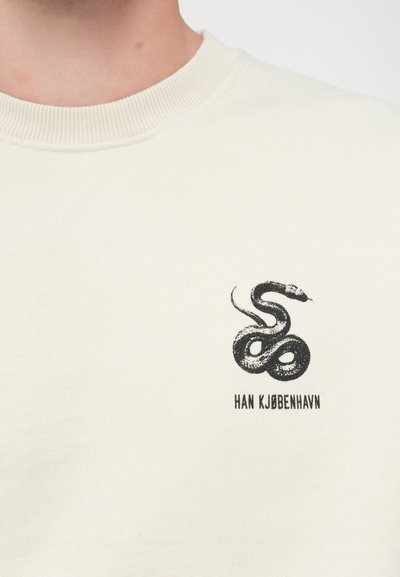 Cremefarvet sweatshirt med et sort slangemotiv og teksten "HAN KJØBENHAVN" nedenunder. Rund ribbet halsudskæring og blød tekstur.