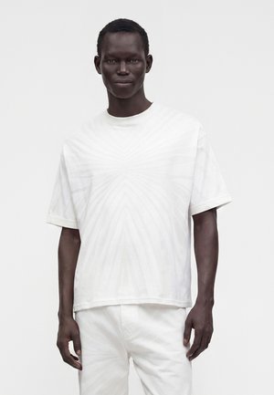 Mann mit dunkler Haut, der ein weißes T-Shirt mit kurzen Ärmeln und weiße Hose trägt und vor einem schlichten hellen Hintergrund steht.
