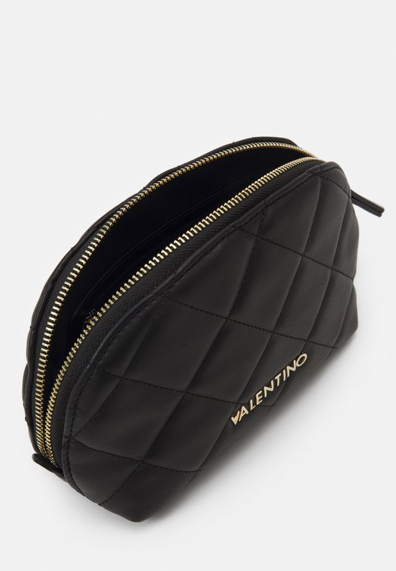 Schwarze Lederkosmetiktasche mit gestepptem Muster, zwei goldenen Reißverschluss-Schließen und eingeprägtem Markennamen in Gold. Glatte Textur und runde Form.
