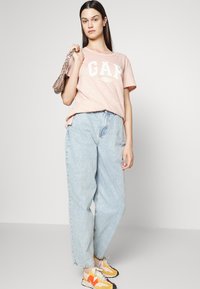 T-shirt de manga curta rosa claro com o logótipo "GAP", combinado com calças de ganga largas azul claro e ténis amarelos com acentos em laranja e preto.