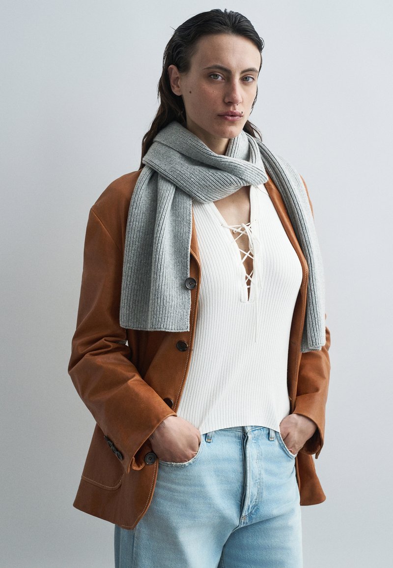 Pelēka, ribota šalle, kas pakārta virs gaiši brūnas ādas žaketes, kas valkāta virs baltas ribotas blūzes un gaiši zilām džinsu biksēm.