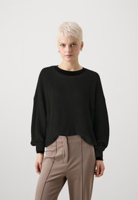 Pulover negru cu nervuri, cu o croială confortabilă, guler rotund și umeri lăsați. Asortat cu pantaloni bej, ajustați, având plisară.