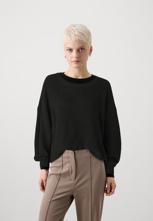Black ribbed sweater with a relaxed fit, round neckline, and dropped shoulders. Paired with beige, tailored pants featuring pleats.