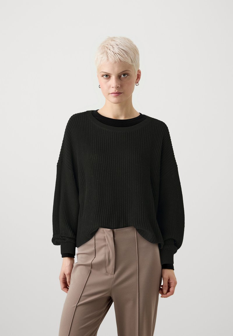 Pulover negru cu nervuri, cu o croială confortabilă, guler rotund și umeri lăsați. Asortat cu pantaloni bej, ajustați, având plisară.