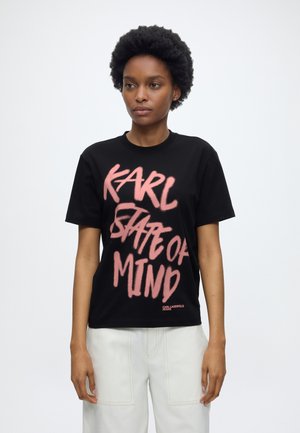 Femme aux cheveux courts et bouclés portant un t-shirt noir avec un texte rose "KARL STATE OF MIND" et un pantalon blanc, debout devant un fond uni.