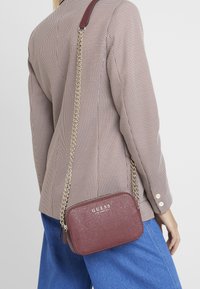 Burgunderfarbene Umhängetasche mit strukturiertem Finish und goldener Kettenriemen, mit GUESS-Logo. Die Tasche hat eine rechteckige Form und einen Reißverschluss.