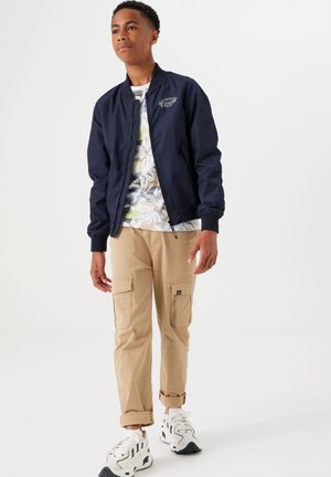 Marine bomberjack met geribbelde manchetten, een wit bloemenhemd eronder, kaki cargobroek met zakken, en witte sneakers met zwarte accenten.
