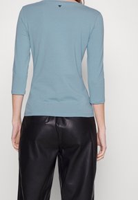 Ljusblå långärmad topp med rundad halsringning, smal passform och subtil logodetalj på baksidan, kombinerad med svarta läderbyxor.