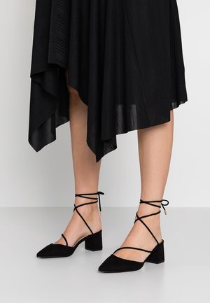 Donna con scarpe nere con tacco a blocchetto e punta affusolata, con cinturini alla caviglia e gonna midi nera plissettata asimmetrica.