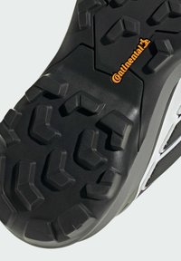 Schwarze Gummisohle mit tiefen Profilen und ausgeprägten sechsseitigen Mustern. Mit orangefarbenem Branding und einem glatten grauen seitlichen Akzent.
