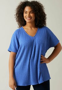 Blå kortærmet top med V-hals, tekstureret stof med zigzag mønster, løs pasform og sideslids for let bevægelse.