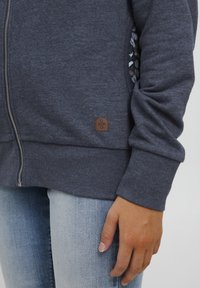 Navy zip-up sweatshirt with a soft texture, featuring a decorative inner lining and a brown logo patch near the hem. Paired with light jeans.