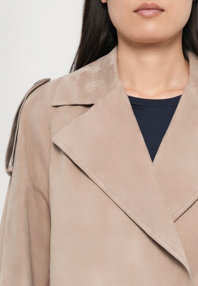 Manteau en daim beige avec un large col, présentant des coutures détaillées et un épaulette unique à l'épaule. Porté au-dessus d'un top navy.