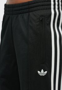 Čierne športové nohavice s elastickým pásom, bočným zipsovým vreckom a tromi bielymi pruhmi pozdĺž nohy. S vyšívaným logom.