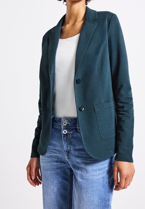 Vrouw draagt een donker teal blazer over een wit topje, gecombineerd met blauwe jeans, staand tegen een effen witte achtergrond.