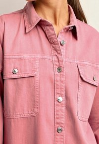 Camisa de mezclilla rosa con cierre de botones en la parte delantera, cuello de punta, dos bolsillos en el pecho y botones de presión metálicos. Textura suave.