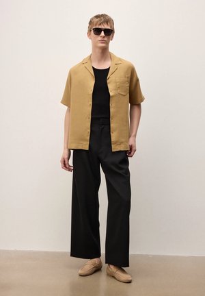 Mladi moški z rjavimi sončnimi očali, oblečen v rumeno srajco s kratkimi rokavi čez črno majico, široke črne hlače in bež copate, stoji.