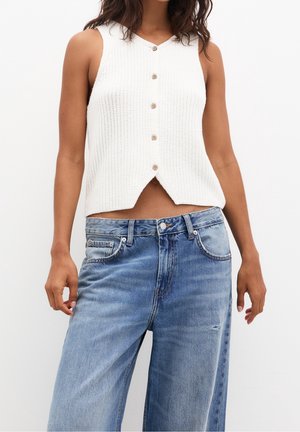 Femme portant un haut blanc sans manches en maille avec des boutons et un jean bleu ample, sur un fond uni.