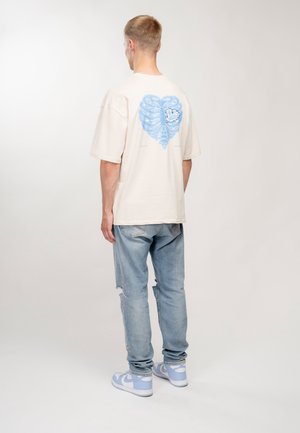 Homme de dos portant un t-shirt ample beige clair avec un motif de cage thoracique bleu, un jean bleu clair déchiré et des baskets blanches et bleues.