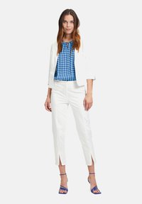 White blazer and pant set over a blue polka dot blouse. Pants feature side slits. Model wears blue strappy heels. Clean, modern design.